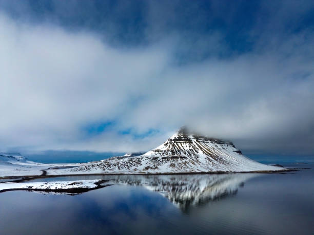 An ethereal drone capture of Iceland's iconic Kirkjufell mountain, partially veiled in low-hanging clouds that lend a mystical atmosphere to the scene. Snow blankets the rugged slopes, creating a stark contrast against the dark, reflective waters below, which mirror the mountain's distinctive shape. The scene exudes a tranquil, almost otherworldly feel, with subtle hints of blue breaking through the cloud cover, offering a glimpse of the sky above. This composition highlights the dramatic beauty and serene isolation of Iceland's winter landscape, making Kirkjufell appear both majestic and serene.