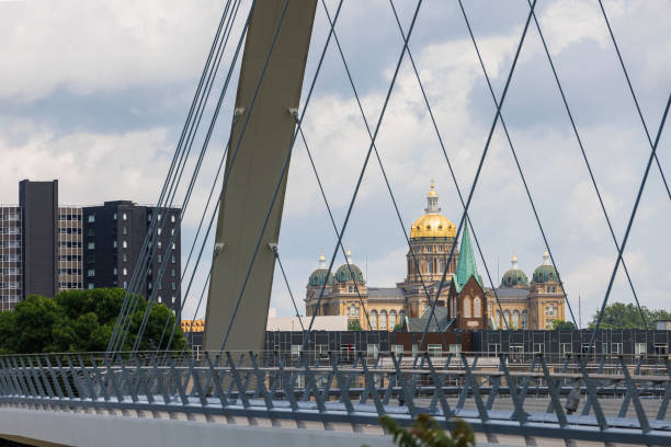 Cable-stayed bridge forms a geometric frame for the Iowa State Capitol, highlighting its golden dome and Renaissance Revival architecture. Steel structure of the bridge contrasts with the ornate 19th-century building, creating a striking juxtaposition in the Des Moines cityscape under cloudy sky