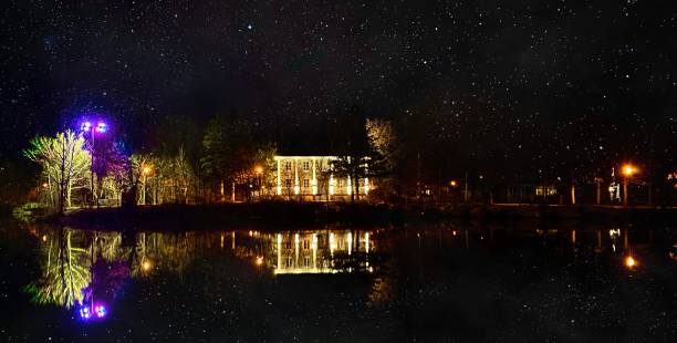 The sky is filled with many stars, and there is a house by the lake, which reflects in the water.