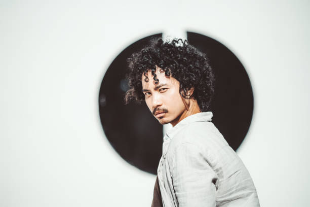 Portrait of a young asian man with curly hair and a mustache, looking confidently at the camera. The subject is dressed in a light, casual shirt with a neutral background