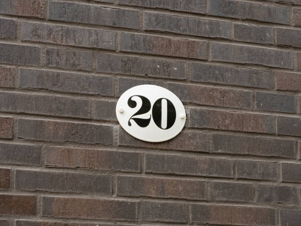 Number 20 on an exterior house wall. The twenty is part of an address. The metal plate is white with black figures. The design is vintage and old fashioned.