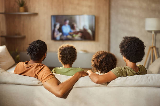 Back view of a black family watching TV at home. Rear view of African American family watching TV on sofa in the living room. couch back stock pictures, royalty-free photos & images
