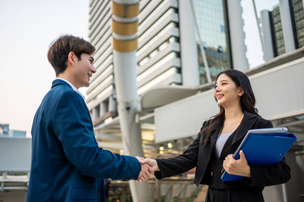 Asian businessman and woman making handshake outdoors in the city. Attractive team of employee people enjoy partnership agreement after negotiations for business deal while standing outside office. hombre y mujer de negocios asiáticos haciendo un apretón de manos al aire libre en la ciudad. el equipo atractivo de empleados disfruta de un acuerdo de asociación después de las negociaciones para el acuerdo comercial mientras está fuera de la oficin