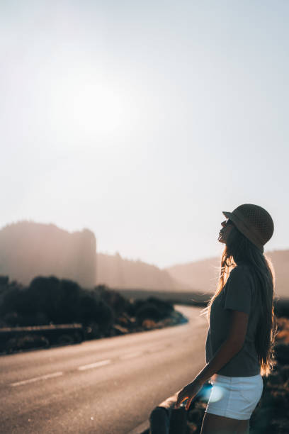 A young woman enjoys the sunny weather, wearing a hat as she gazes up at the bright sky, surrounded by a tranquil landscape and winding road.
