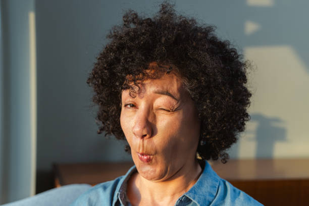 A woman with curly hair makes a humorous face while winking, illuminated by warm sunlight. The playful expression suggests happiness and joy in a casual setting