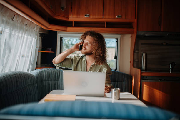 istock Young man working remotely from caravan, making phone call. Man using camper as homeoffice, working on notebook. 2181678393