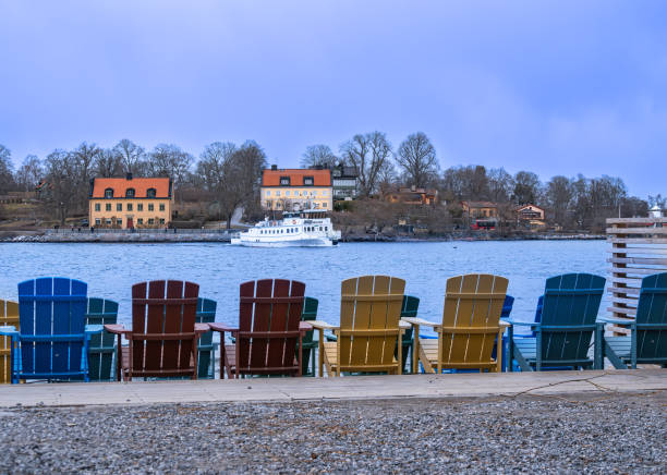 Stockholm, Sweden - 5 March 2023: Colorful Adirondack chairs facing a calm lake with a ferry in the background.