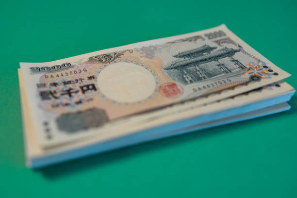 A stack of 2000 yen Japanese banknotes lies on a green background with the top bill showing a temple illustration and security features while the others remain slightly blurred