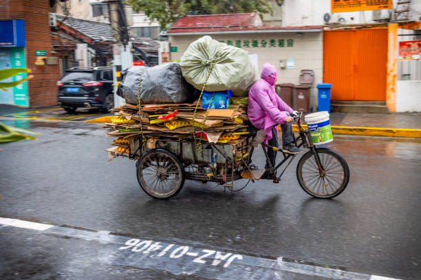 Shanghai, China - 24 Feb 2024: In the midst of a rainy day, a dedicated individual pedals through the streets of Shanghai, transporting an array of recycled materials piled high on a sturdy tricycle, a testament to the city's ongoing efforts in sustainability and resourcefulness.