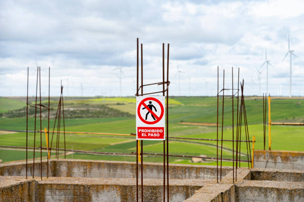 On the iron foundations of an abandoned construction site there is a sign indicating no trespassing, in the background you can see green cereal fields with a field of wind turbines, it is a rural environment.