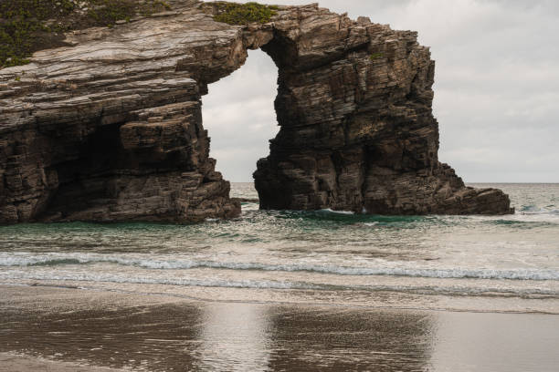 Arch formed by erosion on a large rock located on the beach of LAs Catedrales (Ribade-Lugo) Some small waves reach the beach, leaving residual water and creating a reflection of the rock.