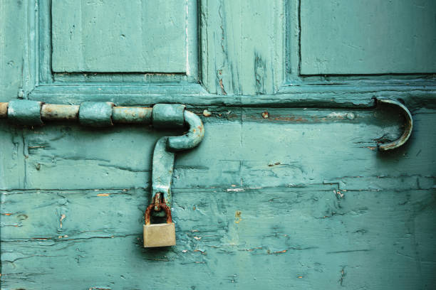 Close-up of a rustic teal-painted wooden door, featuring an old, rusted padlock and latch mechanism. The paint on the door is chipped and worn, revealing layers of age and use. The scene captures the charm of vintage, weather-beaten architecture, with the contrasting textures of metal and wood adding character to the image.