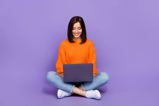istock Full length photo of nice young lady sit floor work laptop coder dressed stylish orange knitted clothes isolated on purple color background 2181724658