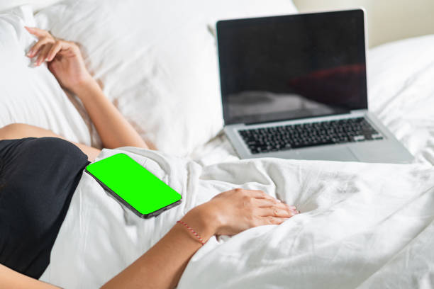 A person lying in bed with a laptop open beside them. A smart phone with a green screen is resting on their stomach. The bedding is white and the setting is cozy and relaxed