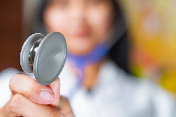 A healthcare professional holding a stethoscope towards the camera, with a blurred background. The person is wearing a white coat, suggesting a medical environment.