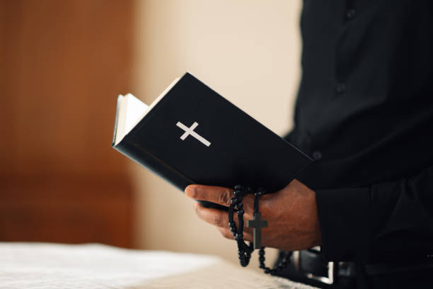 Priest in black attire reads a bible, holding a rosary and cross, showing his deep faith and spirituality