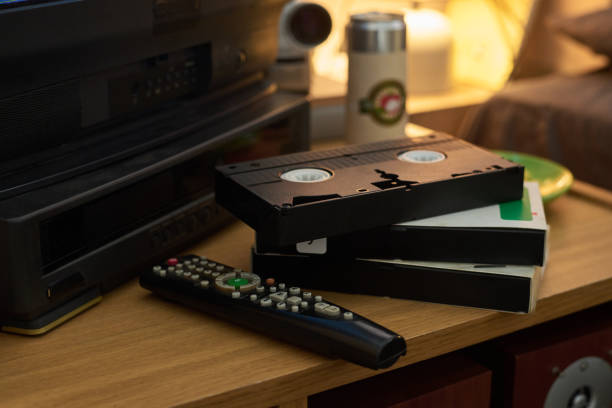Close-up of several vintage VHS tapes stacked near remote control on wooden table with an old TV and other retro items visible in the background. Warm lighting creates nostalgic atmosphere