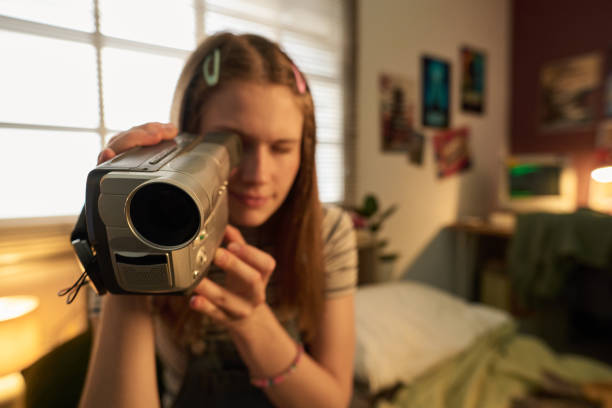 Young woman holding retro handheld video camera in cozy indoor setting with bed and posters in background. Person engaging in creative activity involving digital media