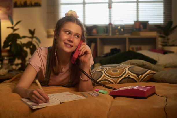 Young woman lying on bed, holding corded retro phone while noting in a journal with relaxed expression. Background showcases cozy bedroom with decorative elements