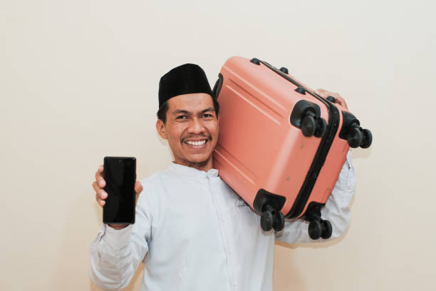 istock Smiling Southeast Asian Muslim man carrying suitcase while showing smartphone screen, Isolated on white background. 2181776287
