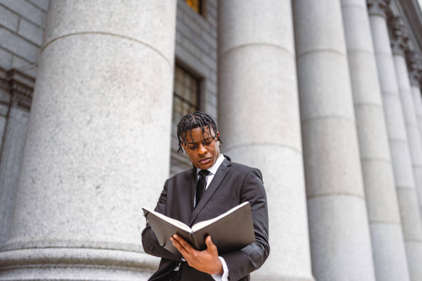 A young professional in a suit reads an open book outside a classical building with large stone columns. The setting suggests a legal or academic context.