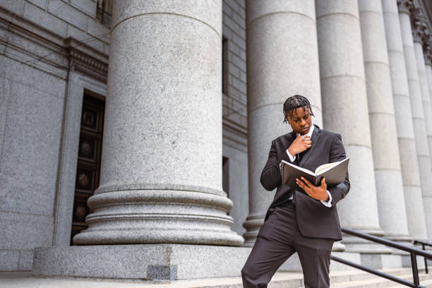 Professional man in business suit reading book and contemplating, standing outside of courthouse with tall columns in background.