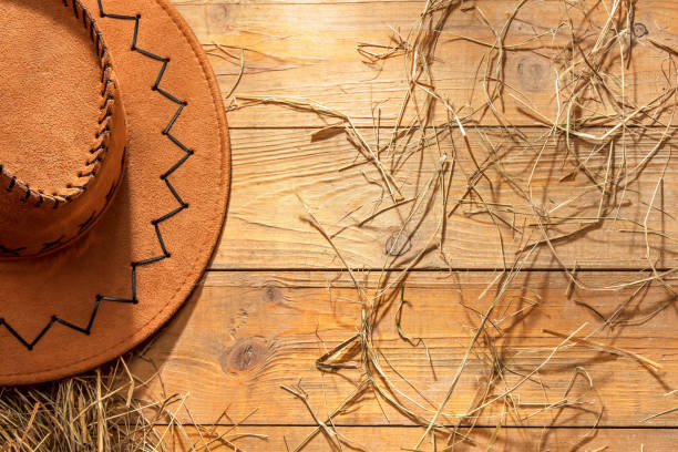 A straw cowboy hat rests on a polished wooden table. In the blurry background, a person is playing an acoustic guitar. The scene evokes a feeling of country music and a rural lifestyle.