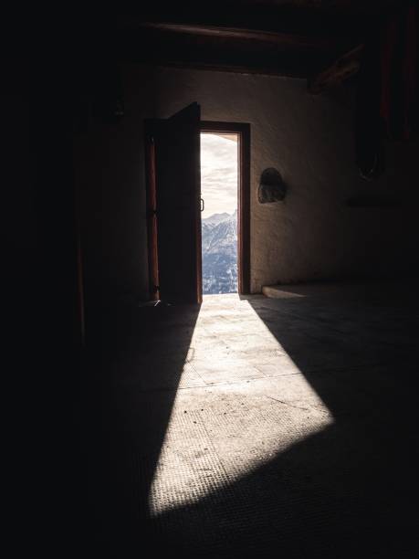 Sunlight casts a long shadow on the cabin floor from an open door that frames a view of distant snow-covered mountains. The interplay of light and shadow creates a tranquil, introspective mood within the rustic interior.