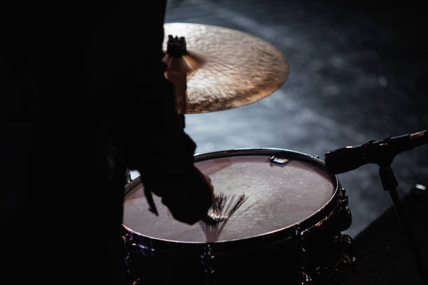 A person is enthusiastically playing a drum set in a dimly lit dark room, creating rhythms that fill the space around them