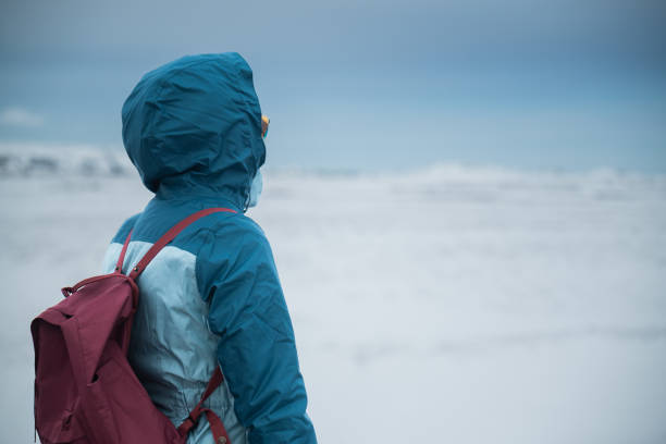 Person bundled up in a blue winter jacket and maroon backpack gazing into a vast, snowy landscape, capturing a sense of solitude and exploration in a winter setting.