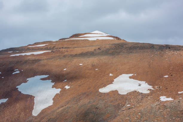 Sunlit snowy pointy peak under gray cloudy sky. Triangle shaped snow mountain top in bright sun. Stony hill of pyramid shape in changeable weather. Sharp rocky snowbound pinnacle in overcast sunny day