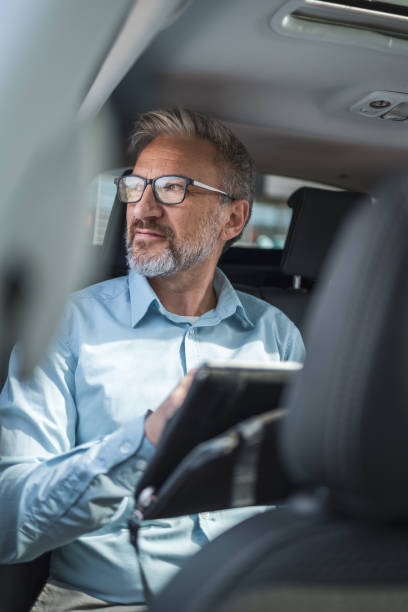 istock Mature businessman riding in a car, working remotely with a tablet. He is dressed in a light blue shirt and looking out the window. He appears focused and thoughtful while commuting. 2181814152