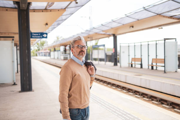 istock Mature man standing patiently at a train station platform, holding his jacket over his shoulder. The setting gives a sense of quiet anticipation and professional readiness, typical of a business commuter. 2181814164