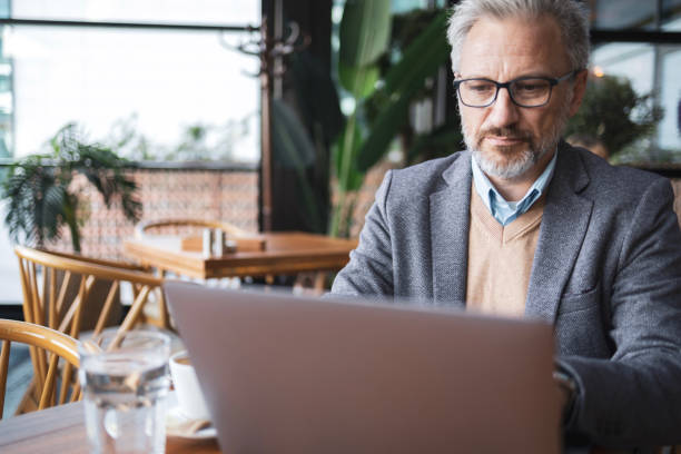 istock Mature businessman working remotely in a cafe. He is focused on his laptop while enjoying a coffee. The setting is modern and comfortable, with natural light and plants. 2181814169