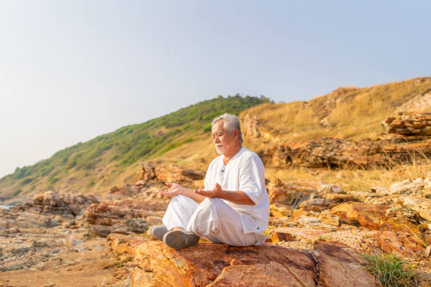 Asian senior man practicing meditation on mountain cliff at sunset.