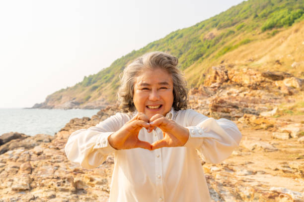 Portrait of Happy Asian senior woman showing hands heart shape during walking at tropical beach at summer sunset. Retired elderly woman enjoy outdoor lifestyle travel nature ocean on holiday vacation.