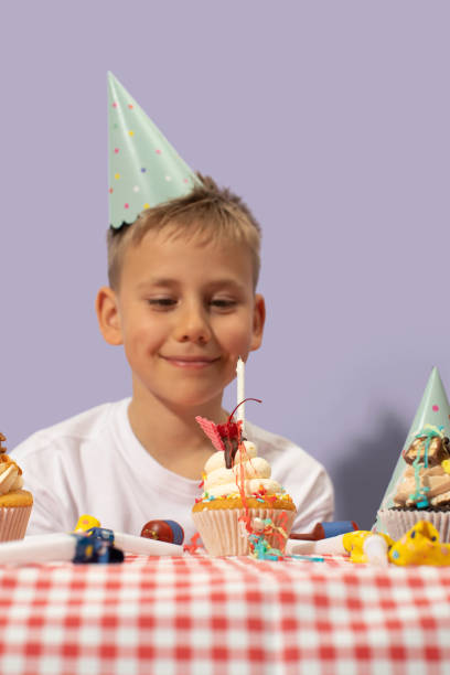 Smiling boy wearing birthday hat and decorated cupcake with lit candle on purple background. Happy child shows homemade dessert with cream over table