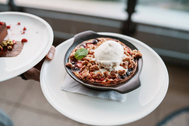 Cropped view of an unrecognizable restaurant server carrying a dish of seasonal berry crisp topped with a scoop of vanilla ice cream to restaurant guests who are seated outside on the restaurant's patio.