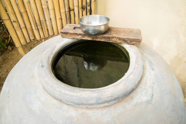 A traditional ceramic water storage jar with a rustic wooden lid and a metal bowl on top, set against a bamboo fence background. This image captures the essence of traditional water storage methods.