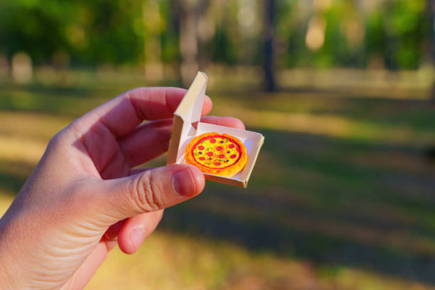 Hand holding a miniature pizza in a small box, set against a blurred outdoor background that suggests a park or forest.