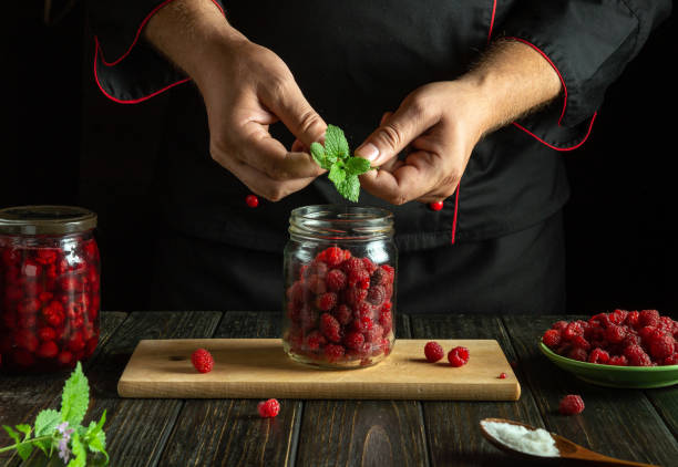 In a cozy kitchen, a chef prepares to infuse fresh raspberries with mint, meticulously layering the ingredients in a glass jar, creating a delightful and aromatic preserve