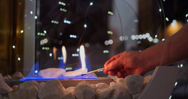 Hand carefully lights biofire with match, bringing biofire soft blue flames to life. Biofire surrounded by stones and gentle bokeh lights creates warm, cozy ambiance perfect for relaxation.