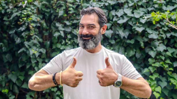 istock A middle-aged, fair-skinned, bearded dark man outdoors in a wooded area, looking at the camera and giving a thumbs-up 2181908432