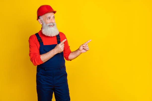 istock Photo of funky positive senior guy dressed uniform overall red hardhat looking pointing empty space isolated yellow color background 2181918531