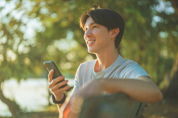 istock Happy young asian man using smartphone while sitting on bench in public park. 2181929072