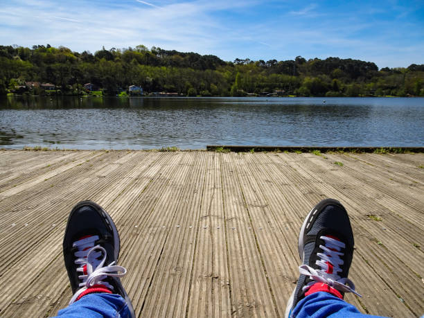 Person wearing jeans and sandals lying on wooden dock by calm lake with mountains in the background. Peaceful outdoor scene with clear blue sky and water. Lakeside relaxation and nature escape.