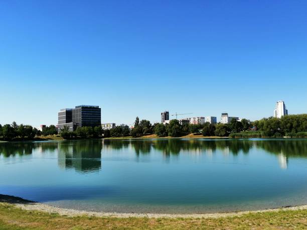 istock A cityscape reflected in a calm lake, with modern buildings, trees, and a clear blue sky on a sunny day 2181947626
