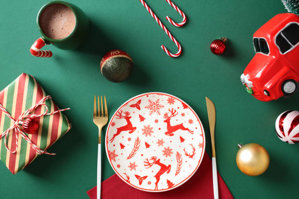 istock Christmas table setting. Red and white reindeer-patterned plate, gold cutlery, and a red toy truck on a green background with Christmas decorations and a cozy hot cocoa mug. Flat lay, top view. 2181954205
