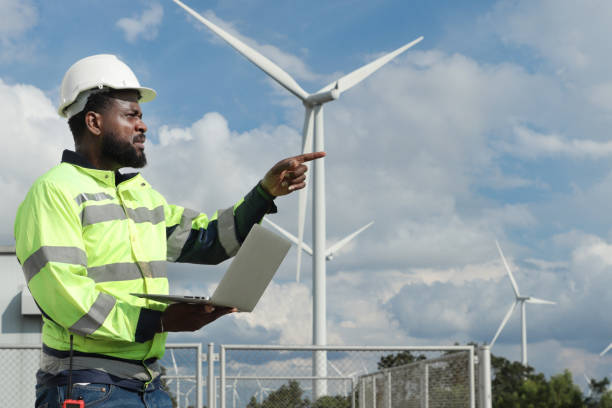 istock African engineer with safety vest and helmet working on laptop computer and pointing at wind turbine farm. Worker works at cloudy sky windmill park. Engineering ecology alternative renewable energy. 2181956669