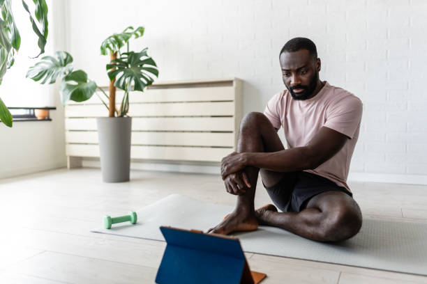 istock Young African-American man sitting on a yoga mat at home, stretching and using a tablet, with small dumbbells nearby. Relaxing in a bright, minimalist room after a workout. 2181961027